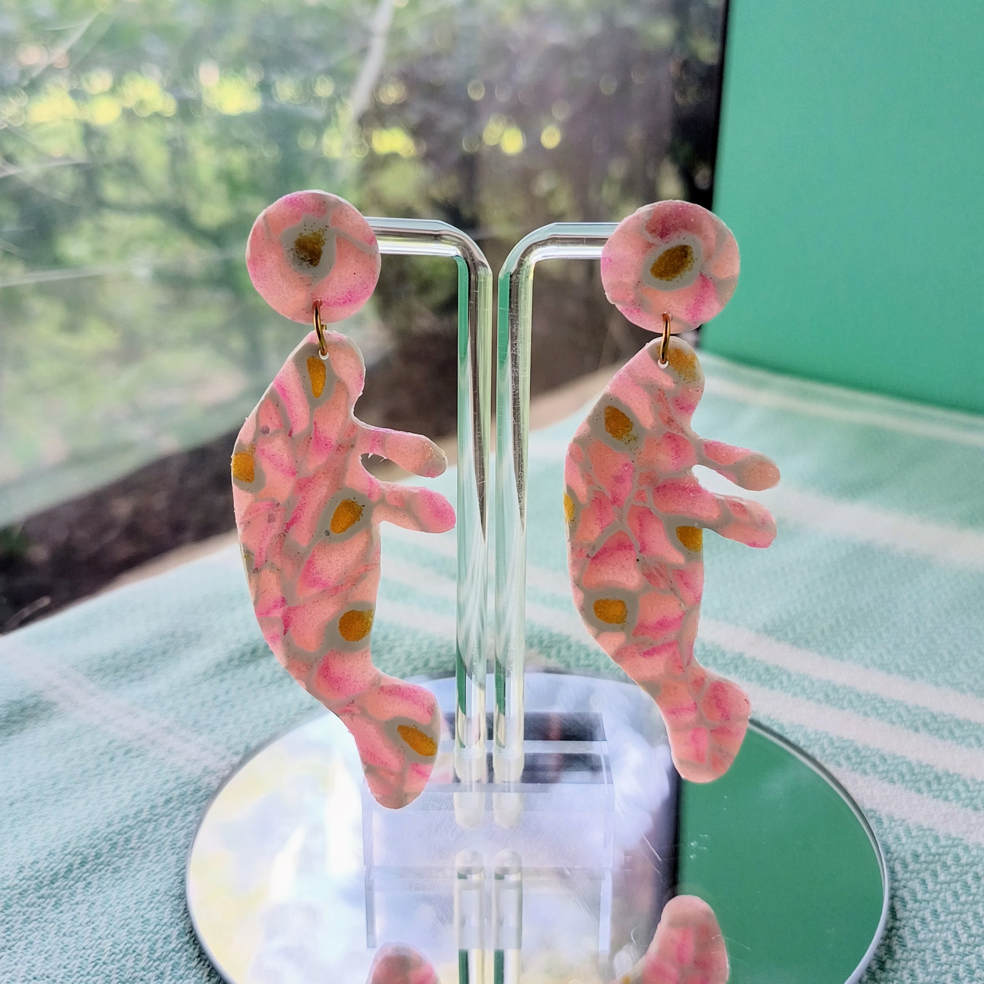 Pink and orange patterned manatee earrings on a clear stand with a blurred green background
