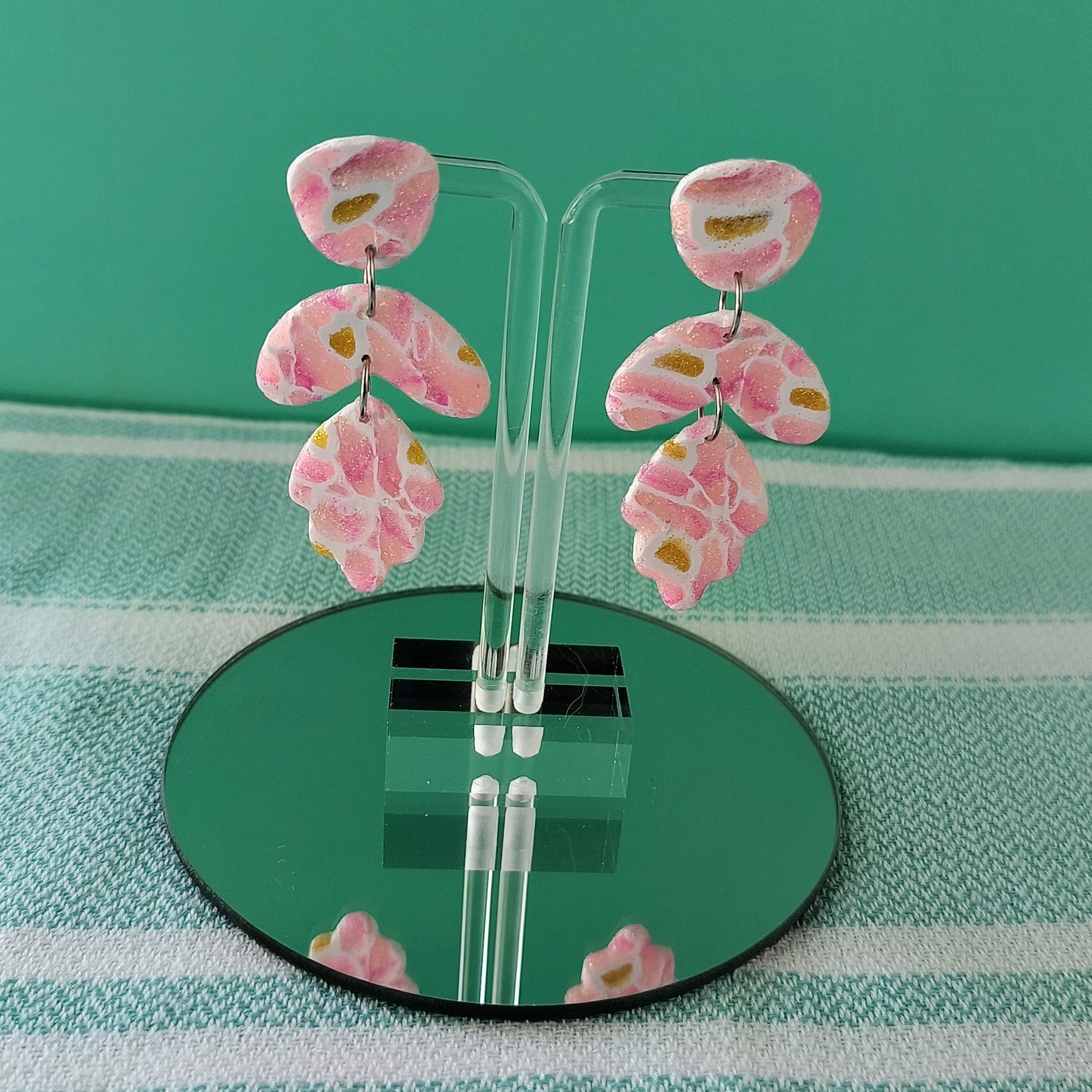dangle polymer clay earrings in sparkle pink, sparkle yellow with white veining, on lucite earring stands, on a circular mirror against a mint backdrop. 