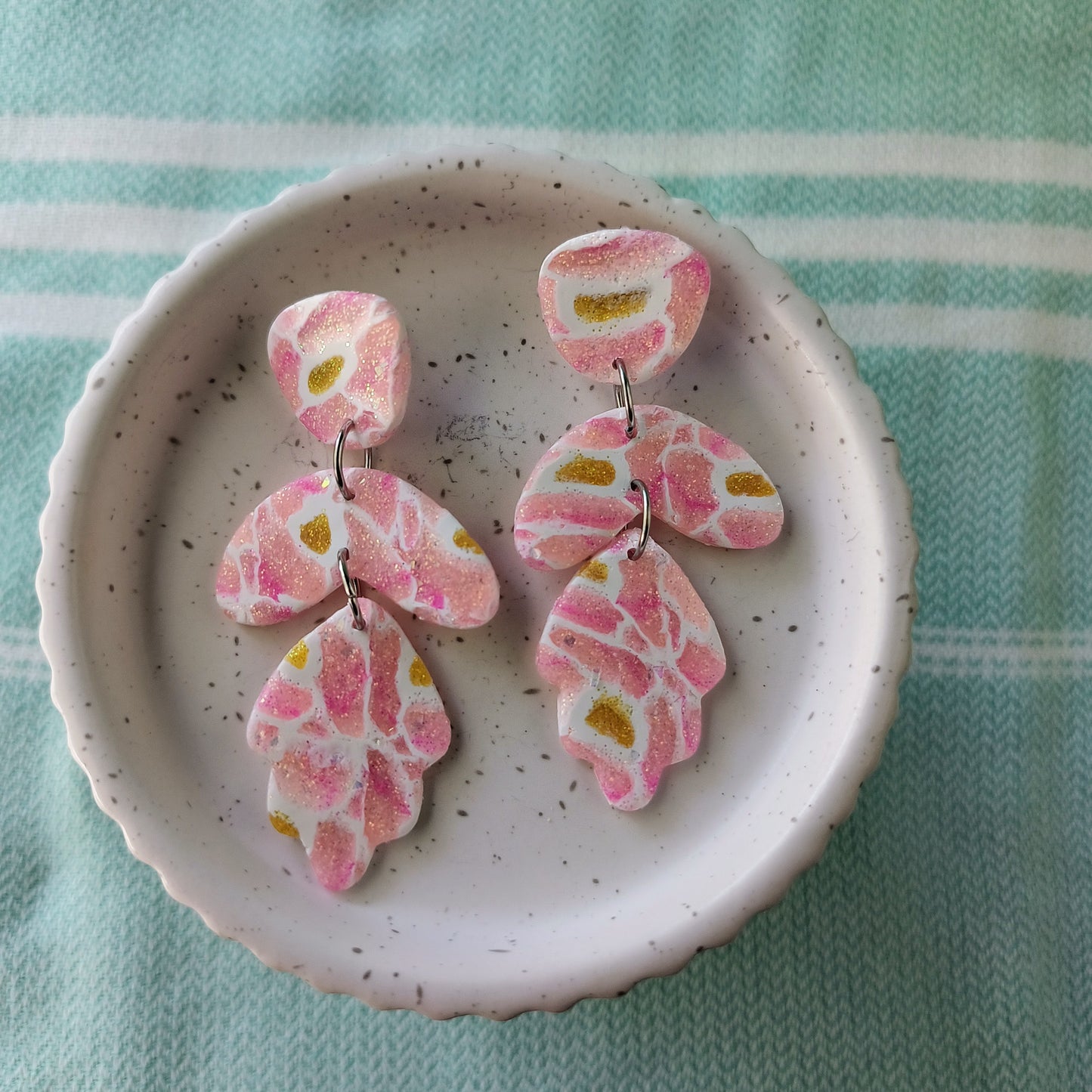 dangle earrings; pink and yellow sparkle polymer clay with white veining, in modern shapes. on small white pedestal on mint striped cloth.