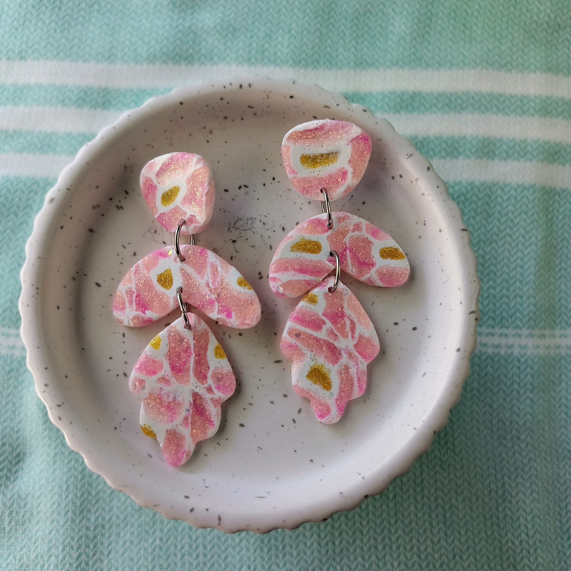 dangle earrings; pink and yellow sparkle polymer clay with white veining, in modern shapes. on small white pedestal on mint striped cloth.