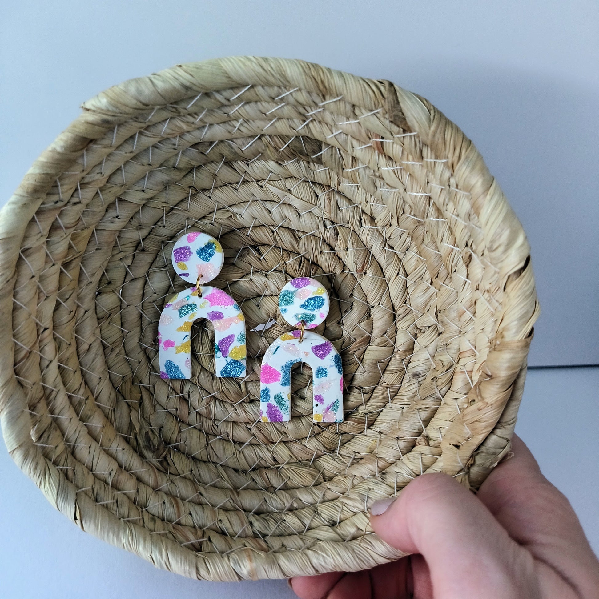 Colorful arch earrings on a woven plate held by a hand.