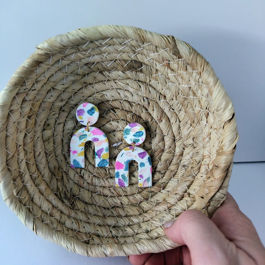 Colorful arch earrings on a woven plate held by a hand.