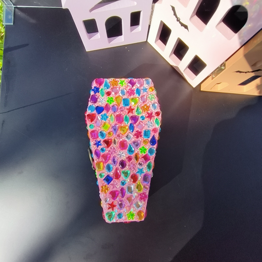 A bedazzled coffin-shaped jewelry box with multicolored gem-like decorations on a black surface.
