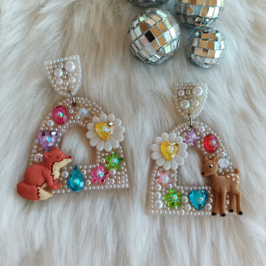 A pair of bedazzled dangle earrings featuring a fawn and fox design with floral, heart, and star motifs, encrusted in shimmering pearls.