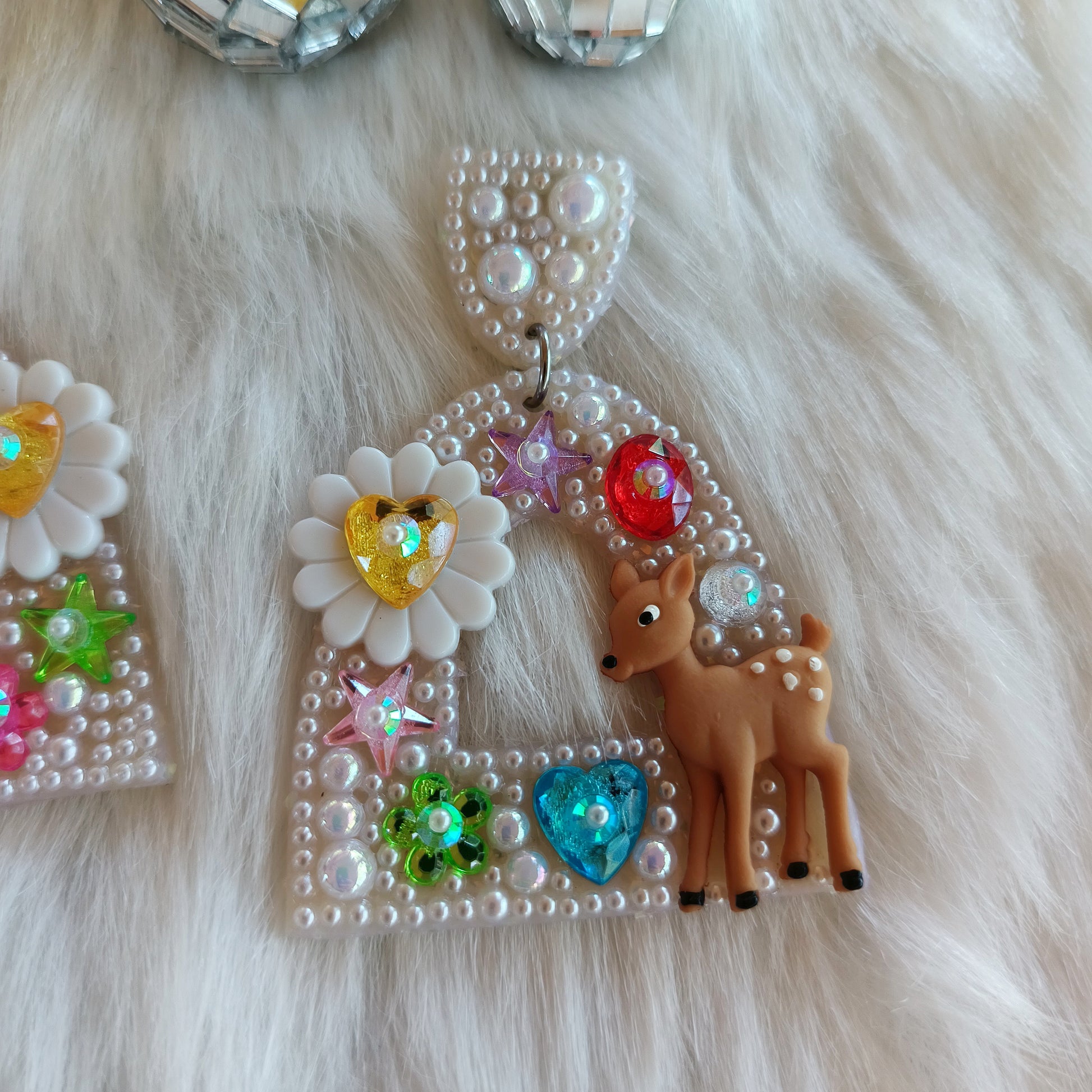 Bedazzled earrings with deer and floral designs on a fluffy white surface.