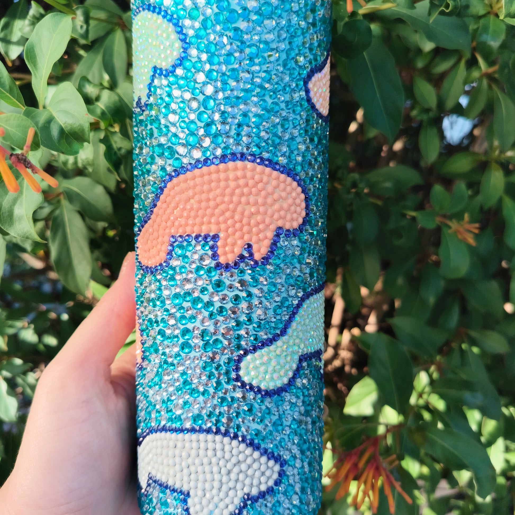 Hand holding a blue bedazzled drink tumbler with manatee design against a green leafy background