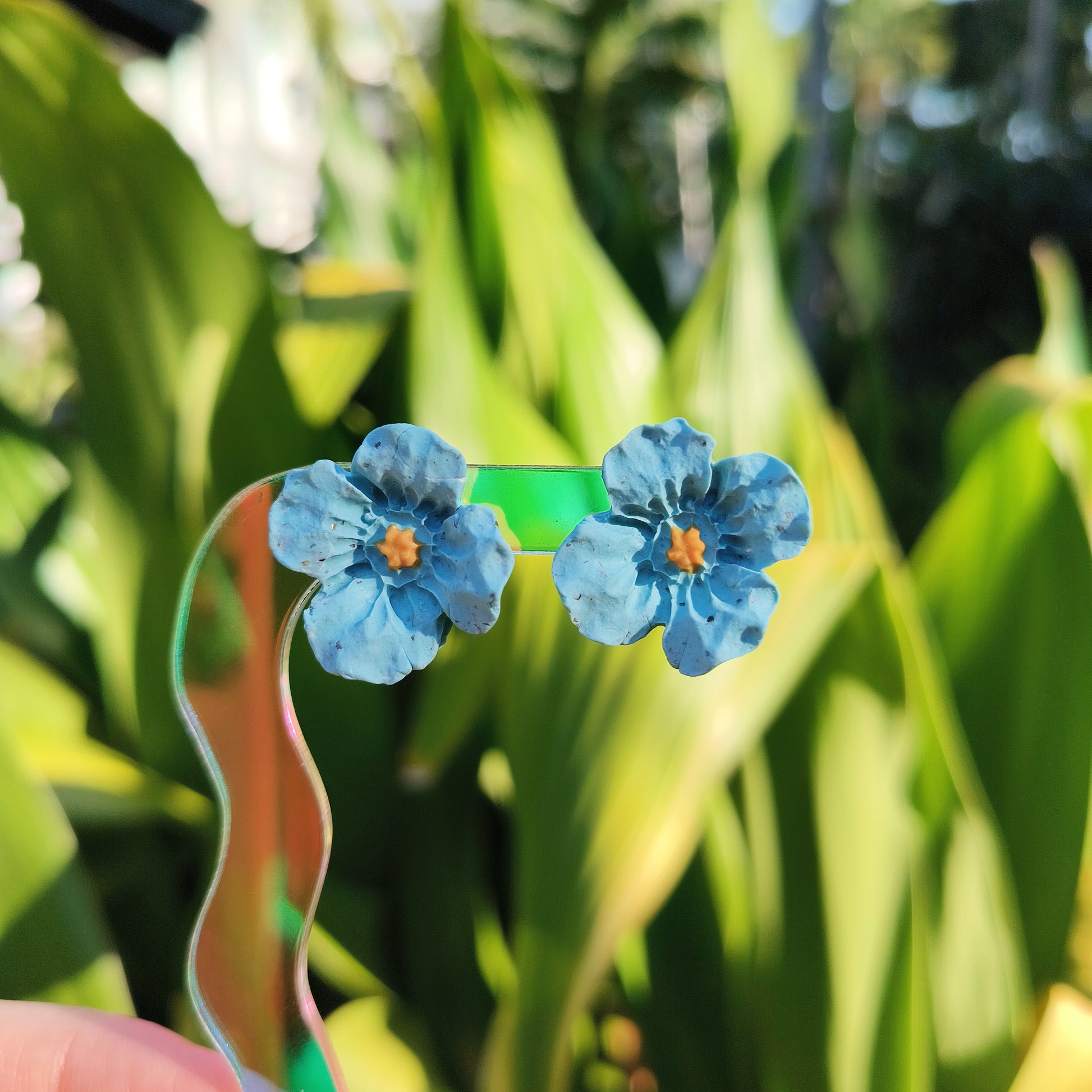 A pair of blue flower stud earrings against a blurred background of greenery.