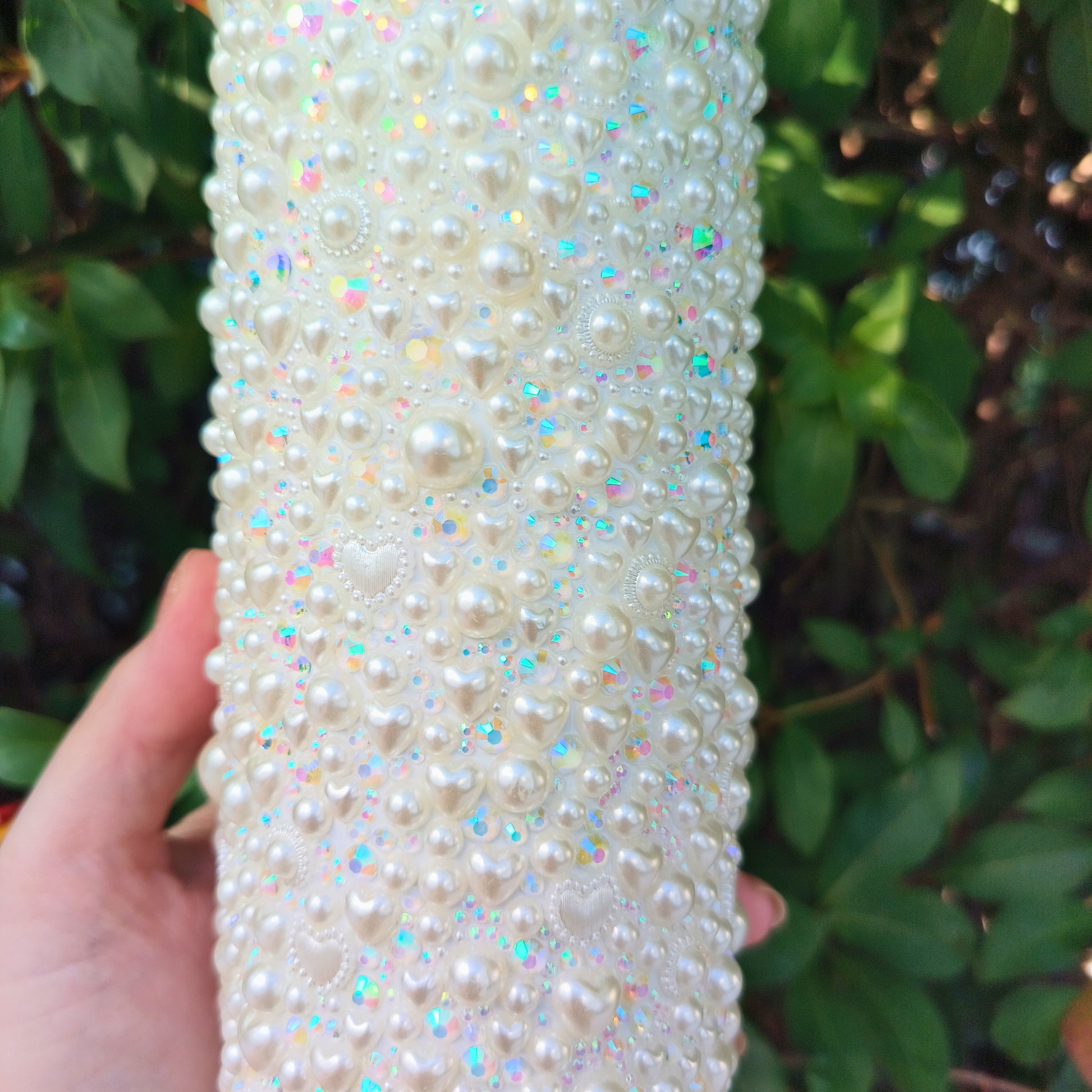 Hand holding a drink tumbler covered in pearls and rhinestones against a green leafy background