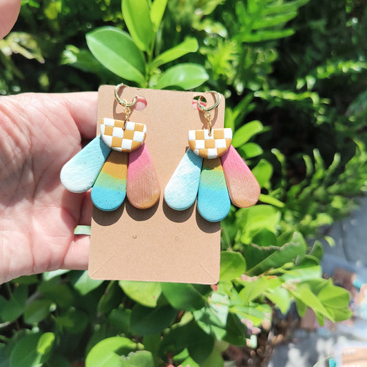 Flower Power dangle earrings with rainbow ombre petals and checkerboard pistol against a backdrop of green foliage.