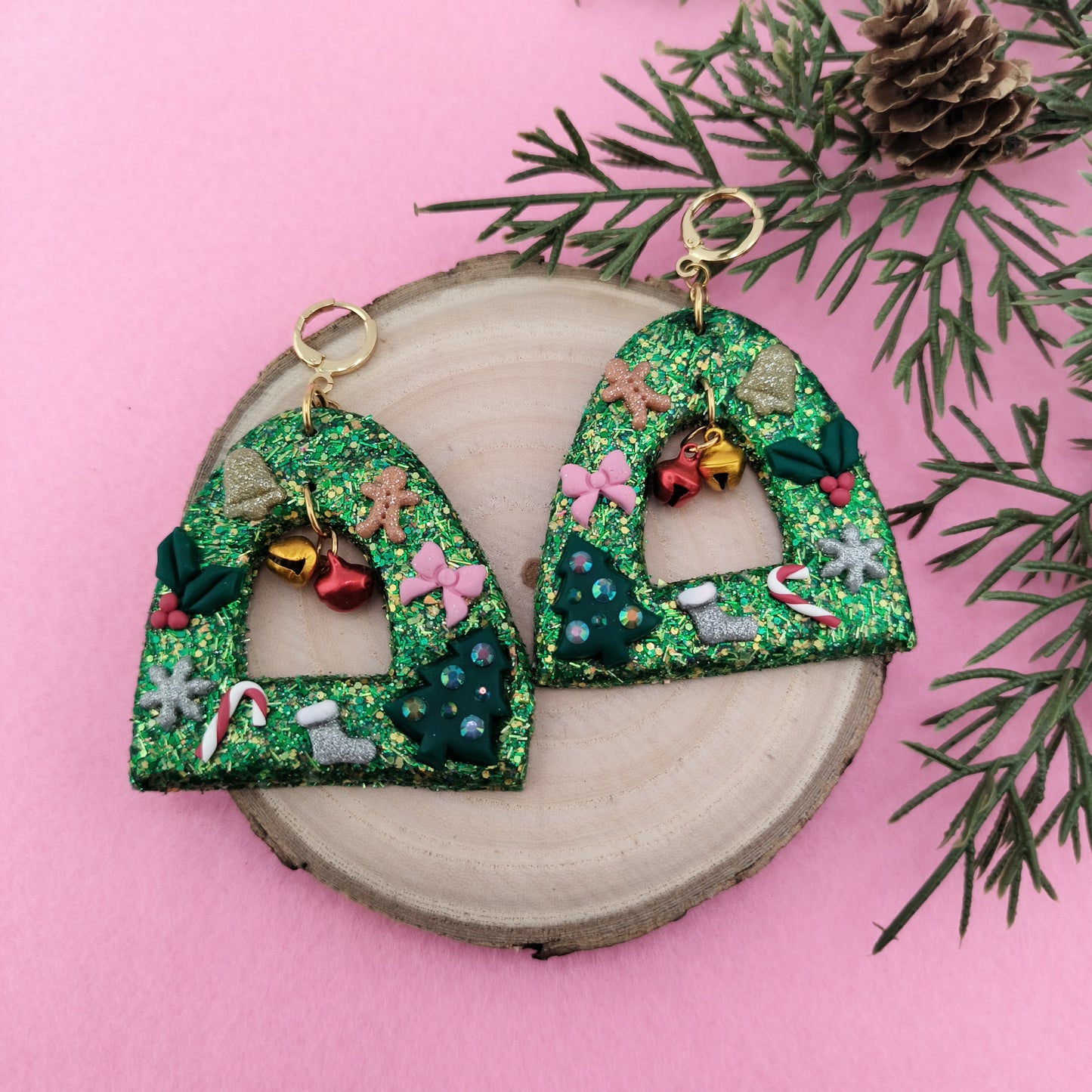 Decorative Christmas earrings with green glittery design on a pink background with greenery.