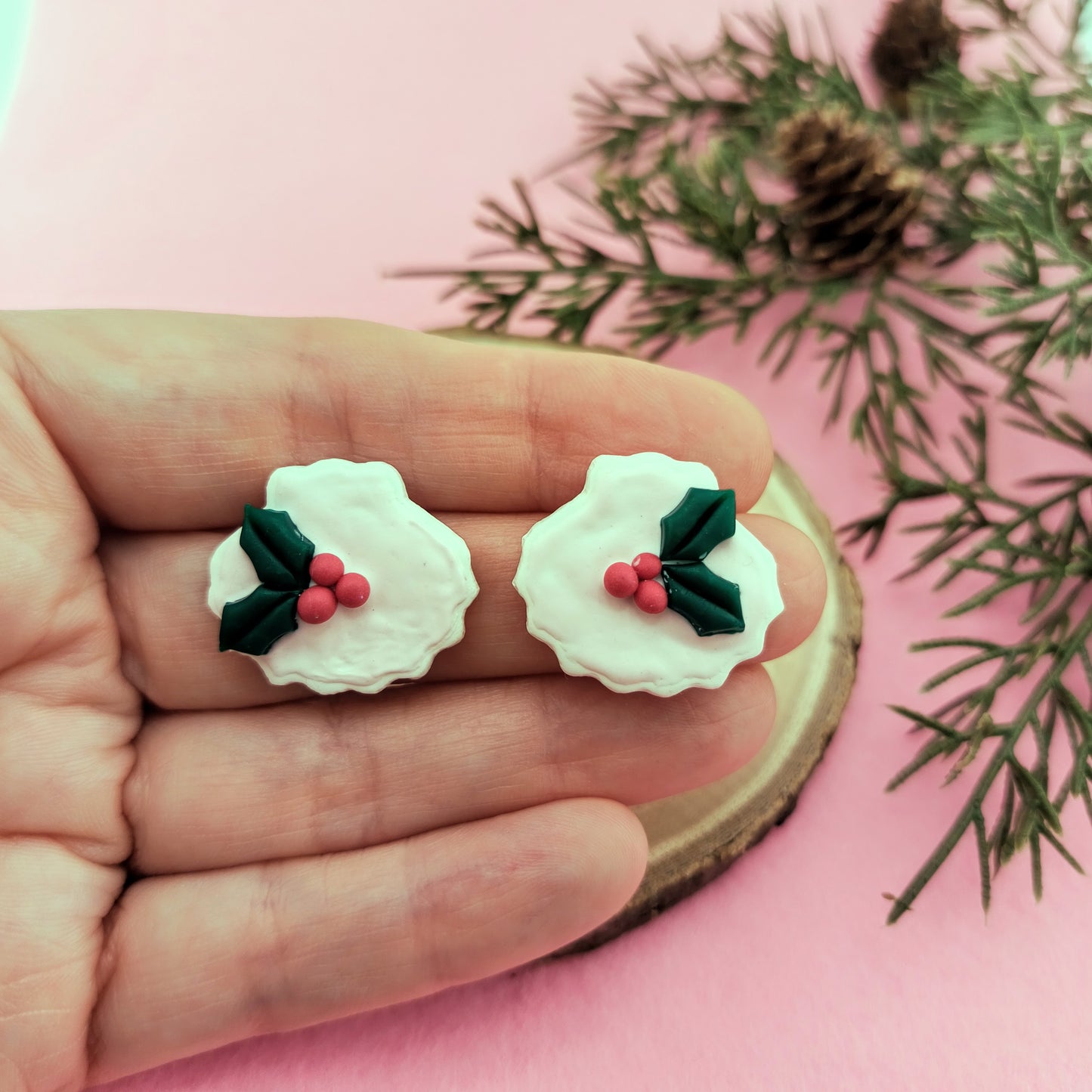 White shell earrings with holly design held between fingers on a pink background with greenery.