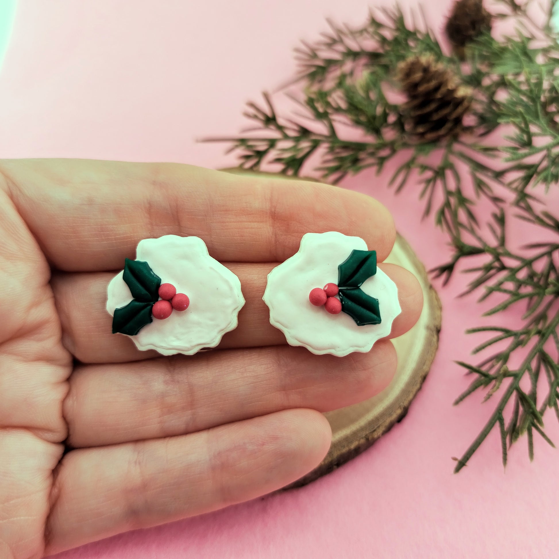 White shell earrings with holly design held between fingers on a pink background with greenery.