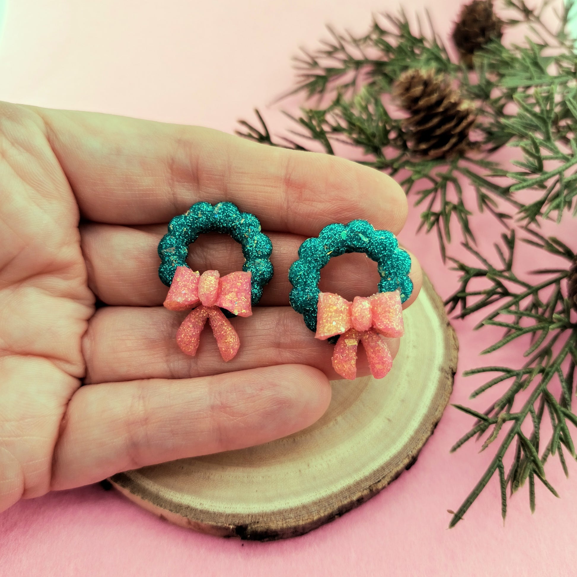 Green wreath earrings held between fingers with a natural wood slice and greenery in the background.