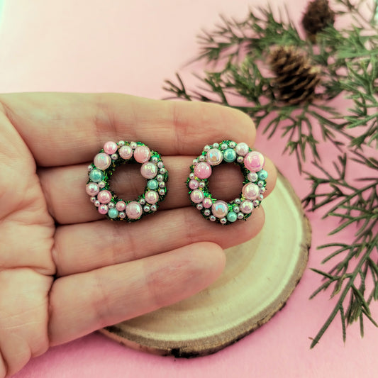 Pair of wreath earrings with pink and green beads held between fingers on a pink background with greenery.