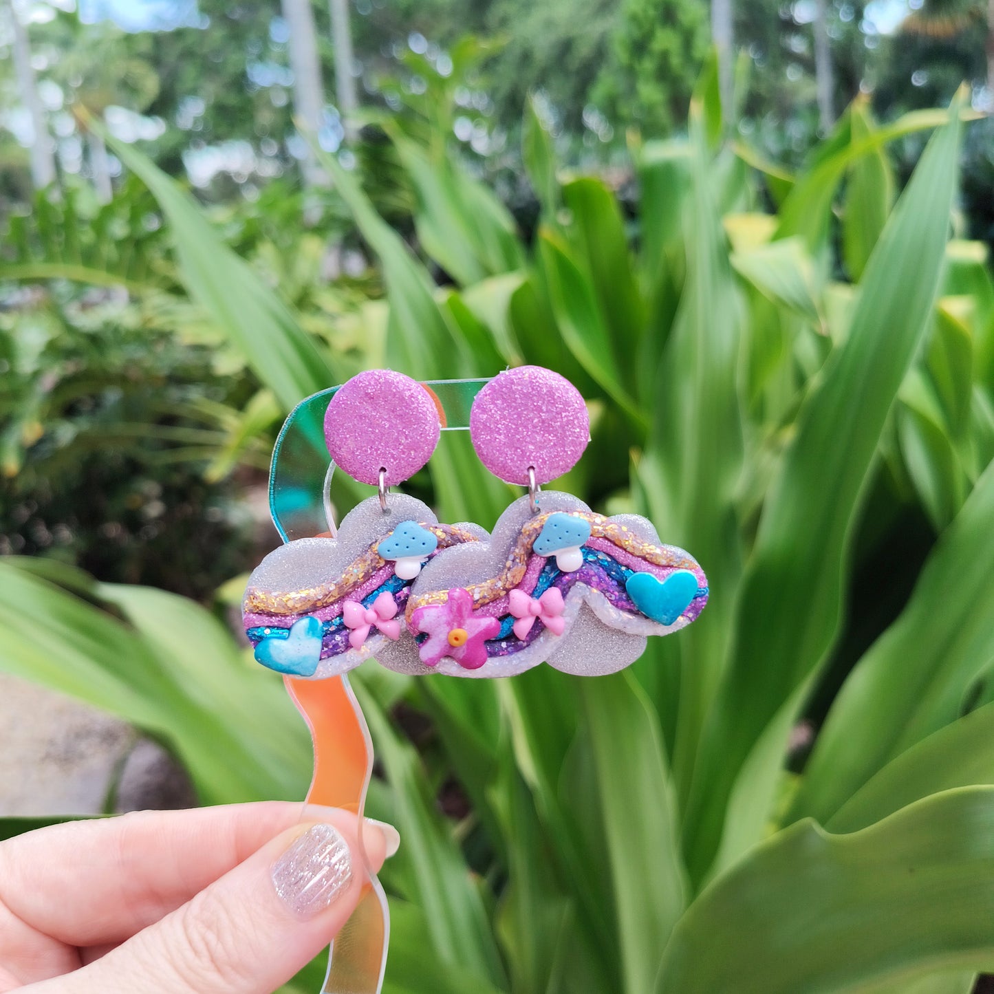 Colorful cloud-shaped earrings held in front of green plants