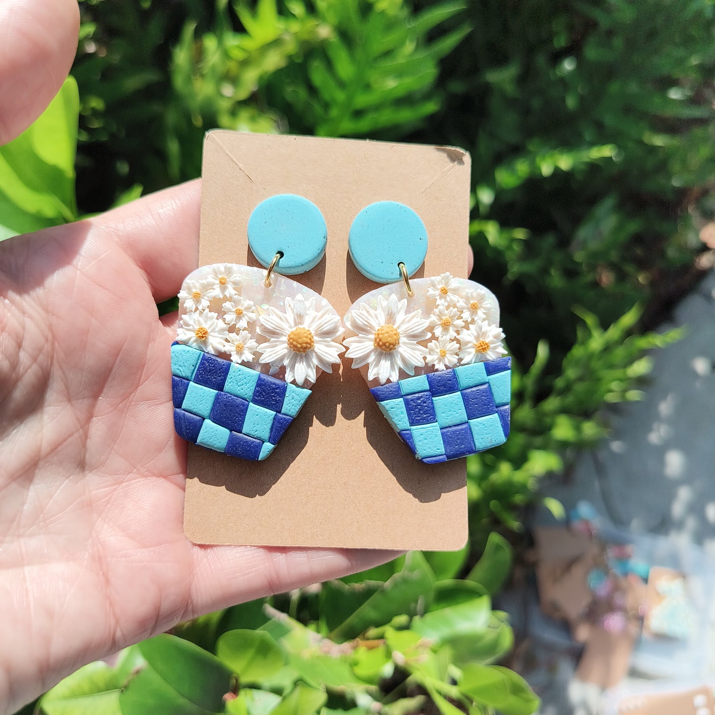 Blue checkerboard baskets holding different sized daises polymer clay dangle earrings, against a backdrop of green foliage. 