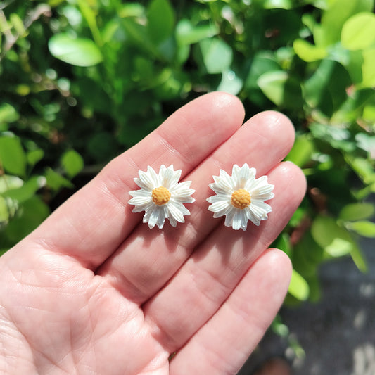 Medium sized white daisy flower stud earrings made from polymer clay, against a backdrop of green foliage. 