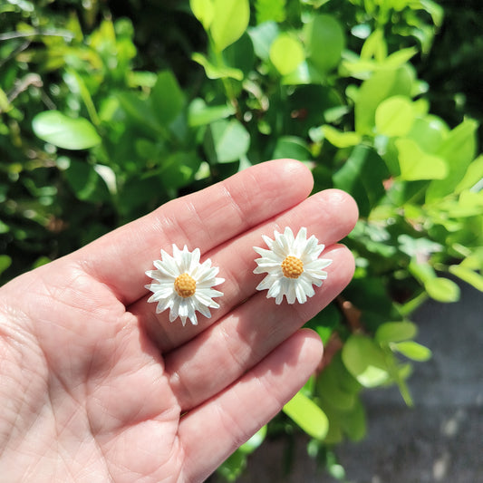 Large sized white clay flower stud earrings against a backdrop of green foliage.