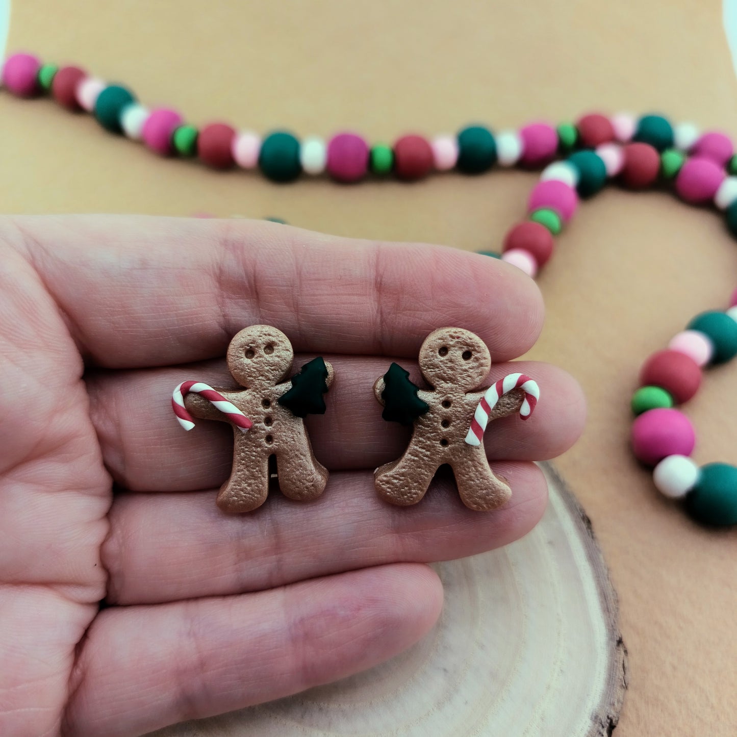 Two gingerbread man earrings with candy canes held between fingers, colorful beaded garland in background.