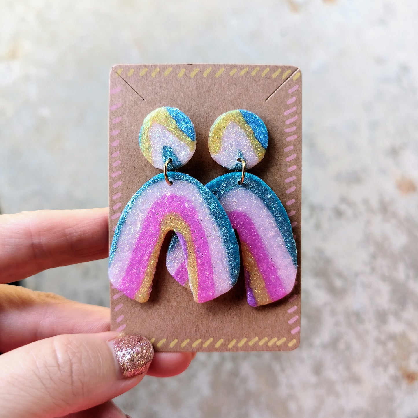 Colorful rainbow-shaped earrings on a cardboard backing held by a hand.