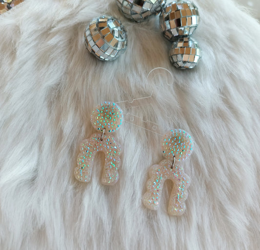 Pair of glittery earrings on a fluffy white surface with disco balls in the background.