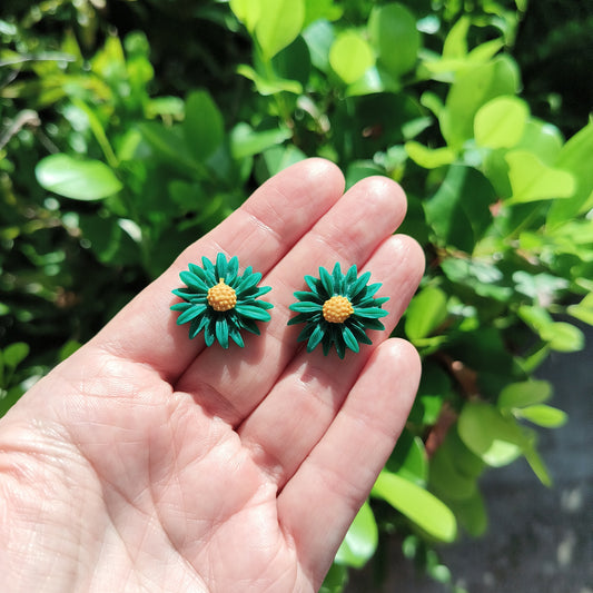 Polymer clay flowers with green petals and yellow pistols stud earrings, against a backdrop of green foliage.