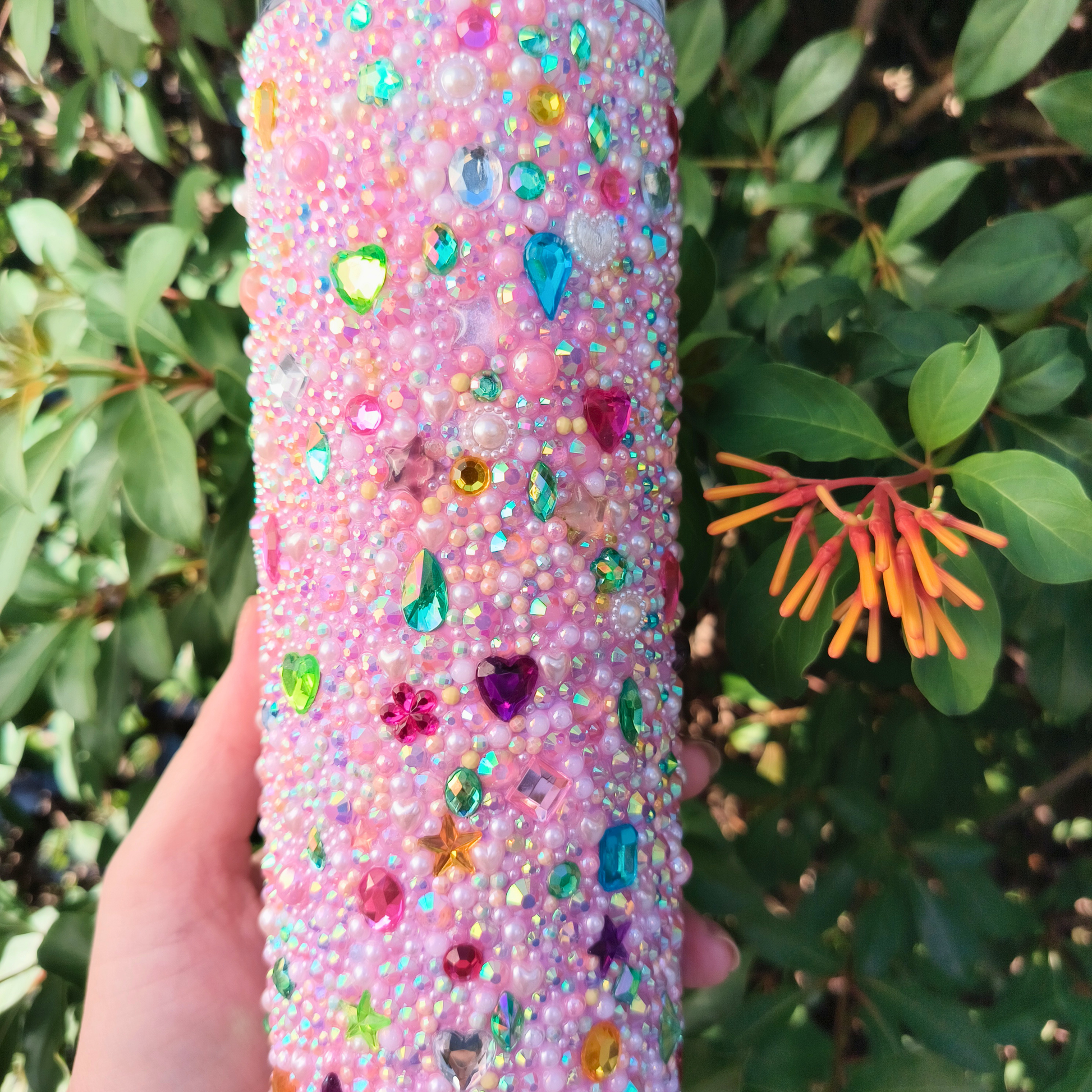 Pink glittery drink tumbler with colorful rhinestones held in front of green foliage