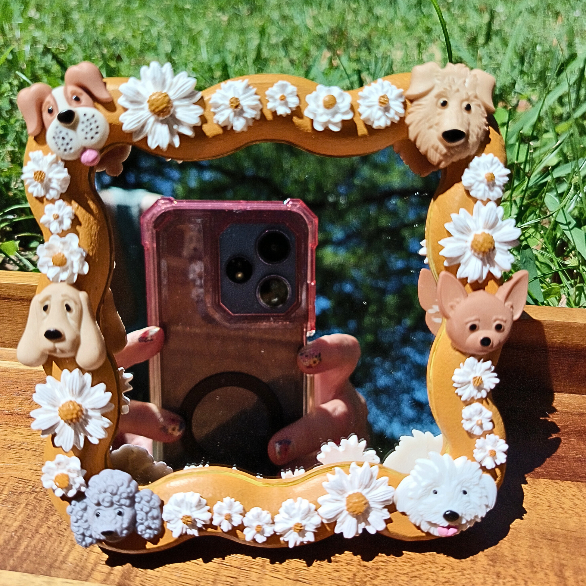 A decorative mirror with a wooden frame featuring polymer clay daisies and dog shapes, displayed outdoors with a smartphone reflecting in the mirror.
