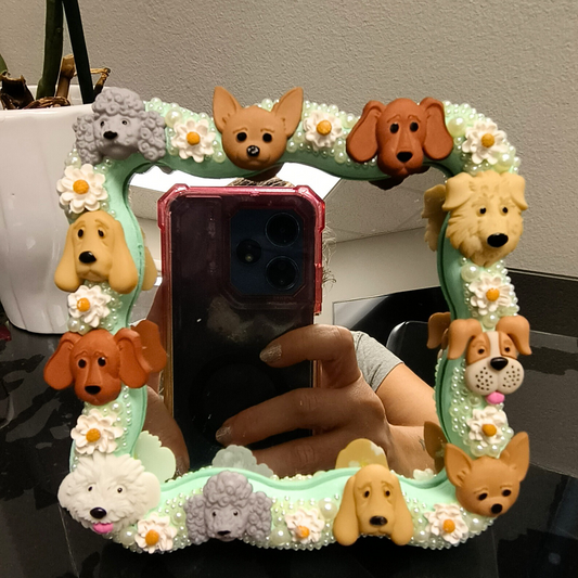 A decorative tabletop mirror with a frame made from wood decorated with dogs, daisies, and pearls. The mirror is displaying a mobile phone reflection and is placed on a desk.