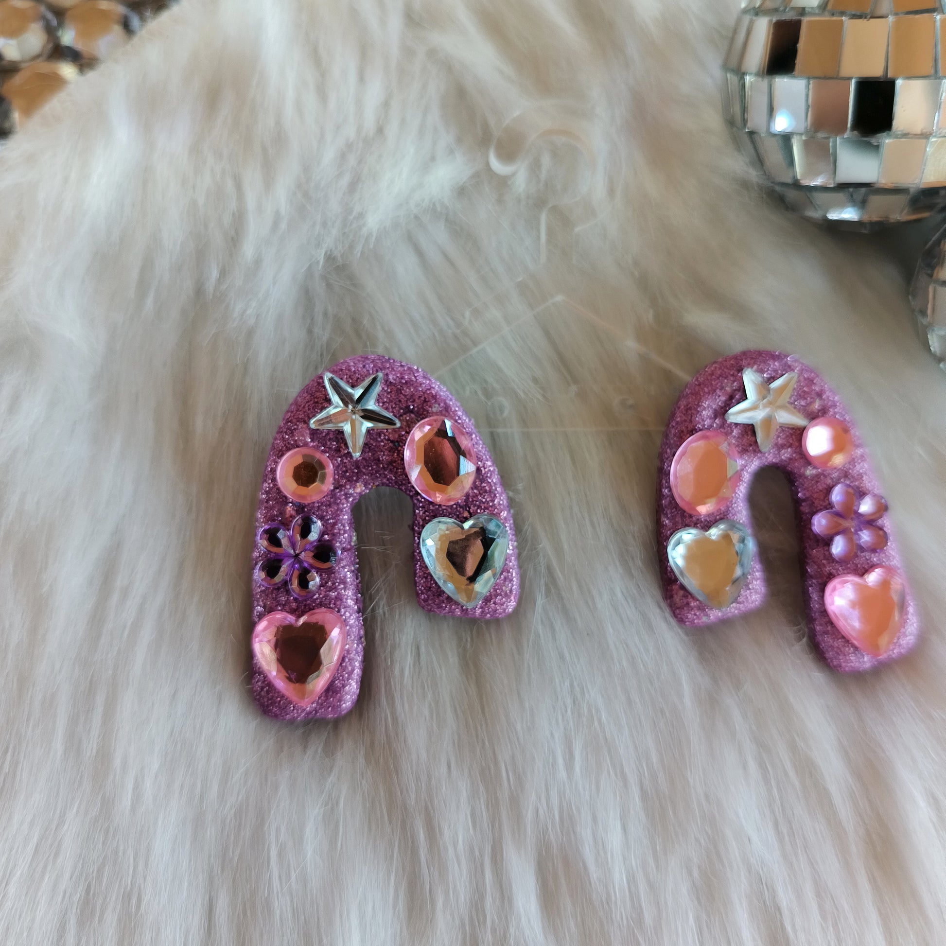 Two pink sparkle stud earrings with hearts and stars on a fluffy white surface.