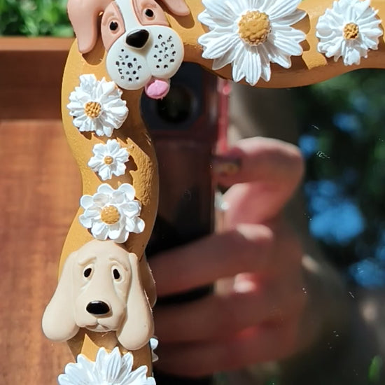 A decorative mirror with a wooden frame featuring polymer clay daisies and dog shapes, displayed outdoors with a smartphone reflecting in the mirror.