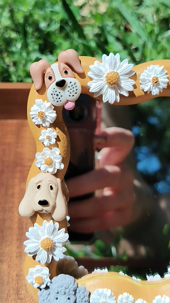 A decorative mirror with a wooden frame featuring polymer clay daisies and dog shapes, displayed outdoors with a smartphone reflecting in the mirror.