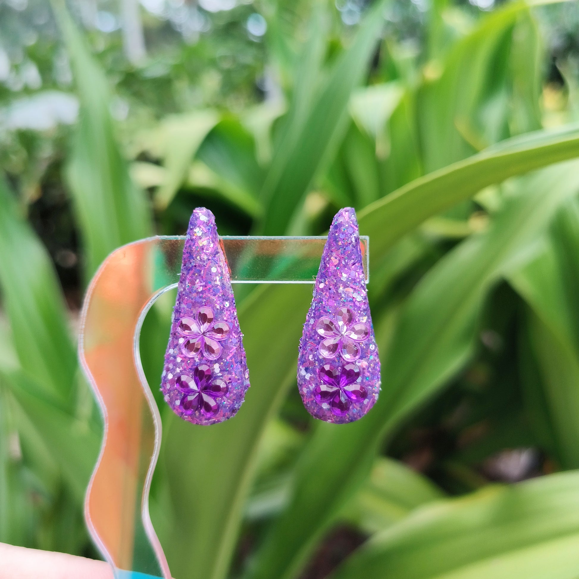 A pair of purple teardrop-shaped earrings with a sparkling design, displayed against a blurred background of greenery.