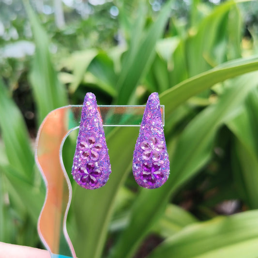 A pair of purple teardrop-shaped earrings with a sparkling design, displayed against a blurred background of greenery.