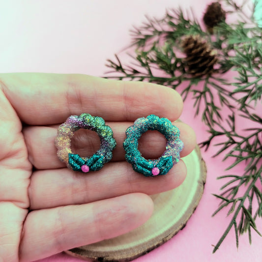 Colorful wreath-shaped earrings held in a hand with a pink background and greenery.