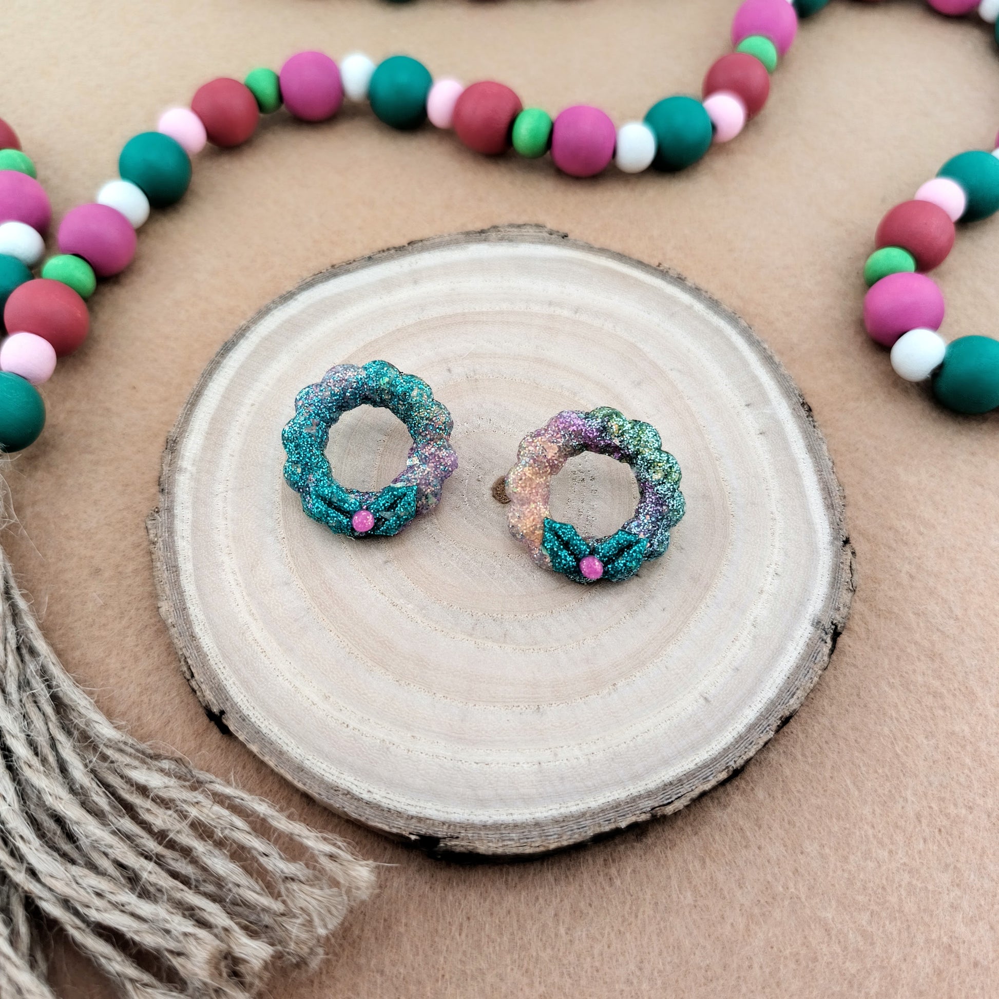 Colorful wreath shaped earrings on a wood slice with a beaded garland in the background/