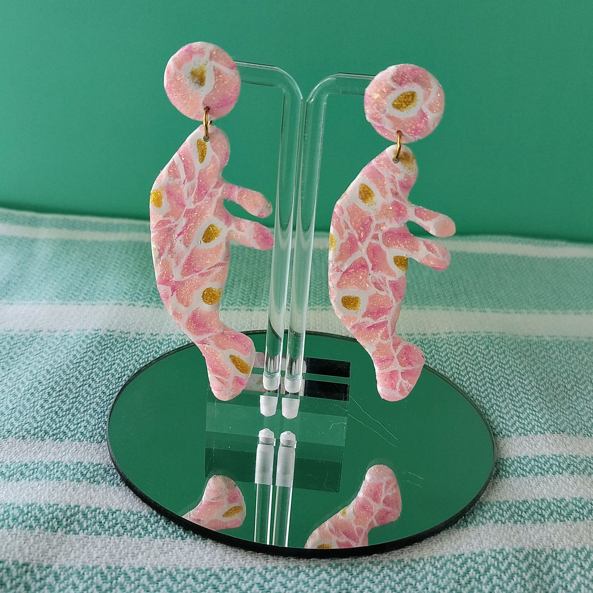 Pink, yellow, and white manatee earrings on a clear stand against a green and white striped background