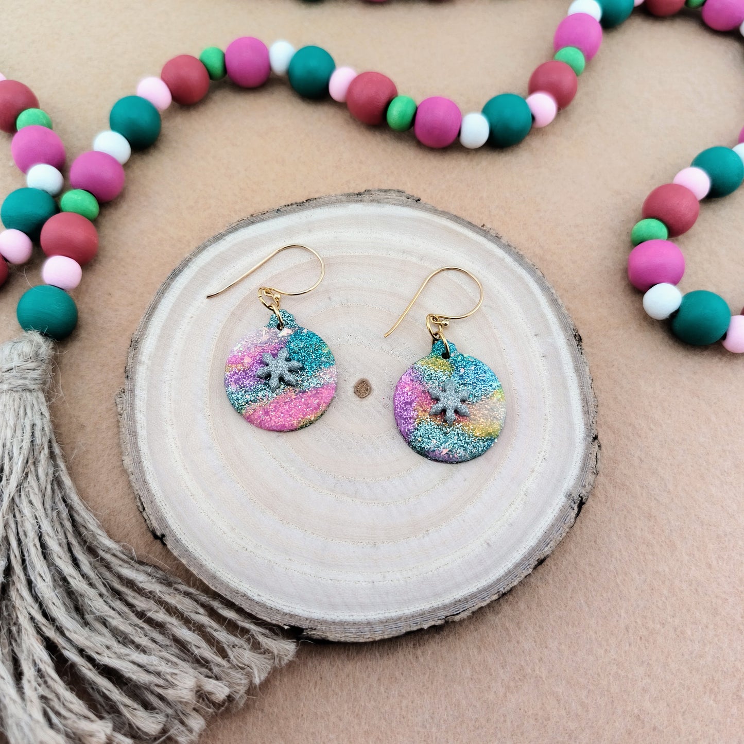 colorful ornament earrings with snowflake detail displayed on a wood slice with a beaded garland in the background