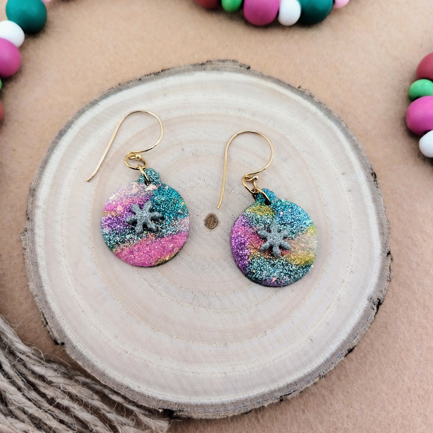 Colorful sparkle snowflake ornament earrings on a wooden surface with a beaded garland in the background