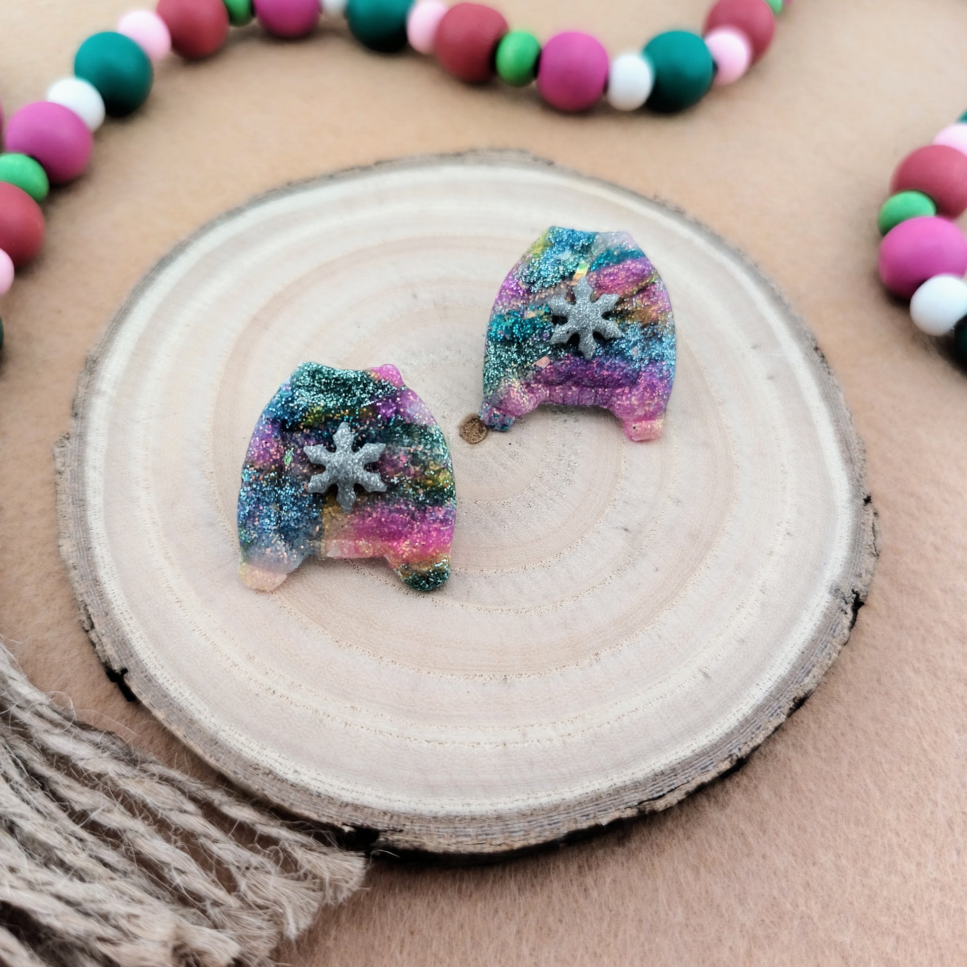 Colorful glittery sweater shaped earrings on a wooden surface with a beaded garland in the background.
