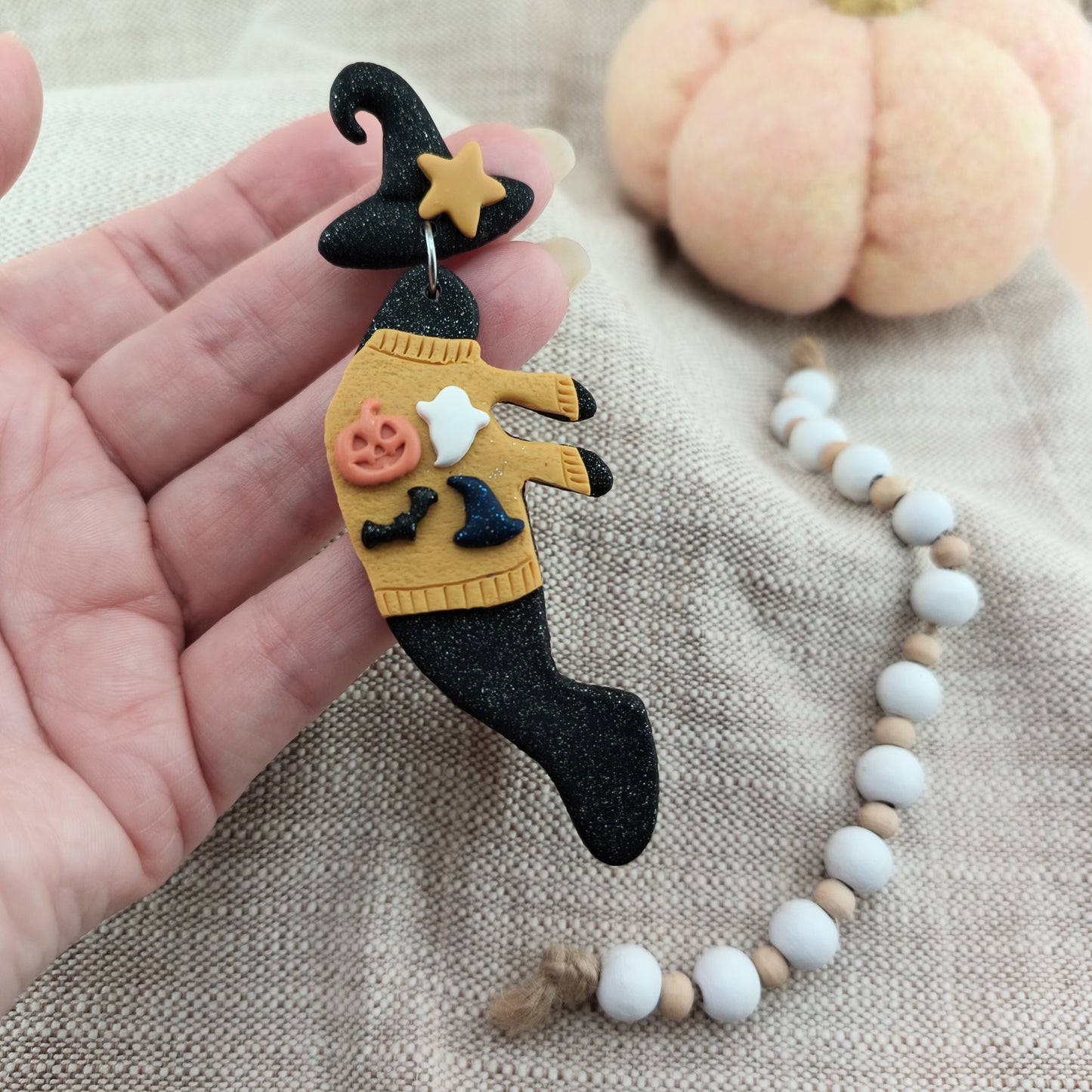 Halloween-themed manatee earring with a witch design held by a hand, with a pumpkin and beaded necklace in the background.