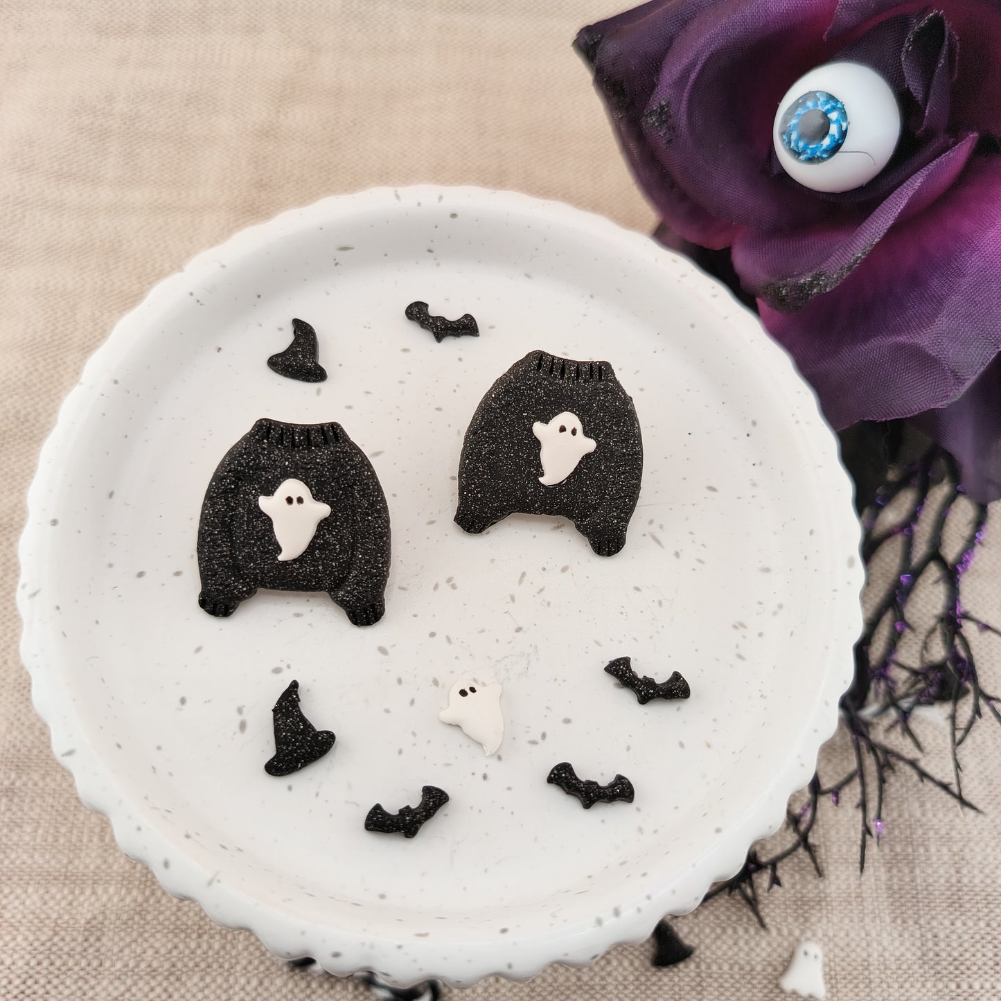 White dish with stud earrings featuring bats on a beige surface with a purple flower.