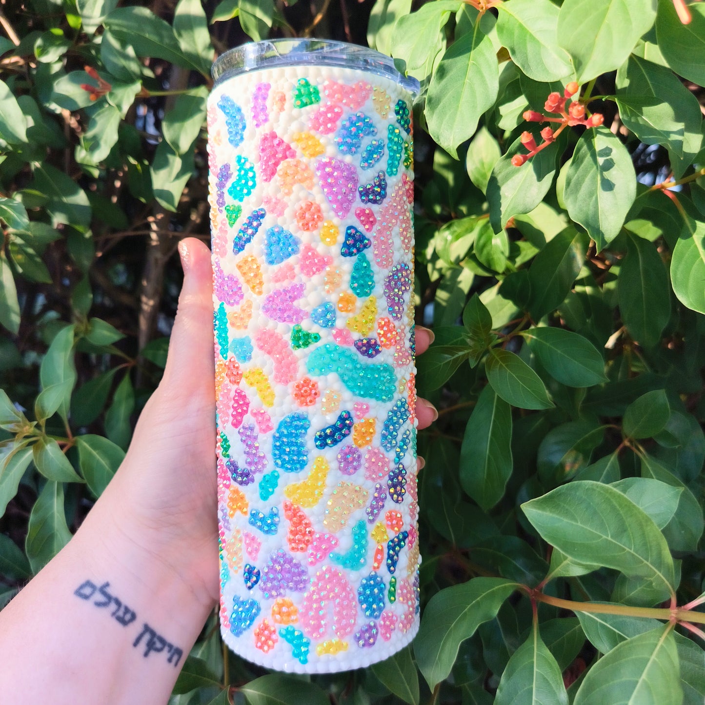 Colorful bedazzled tumbler held in front of green foliage