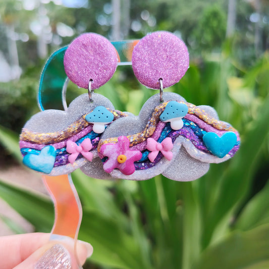 A pair of handcrafted dangle earrings with multicolor sparkle clay designs, resembling clouds, displayed against a plant backdrop.
