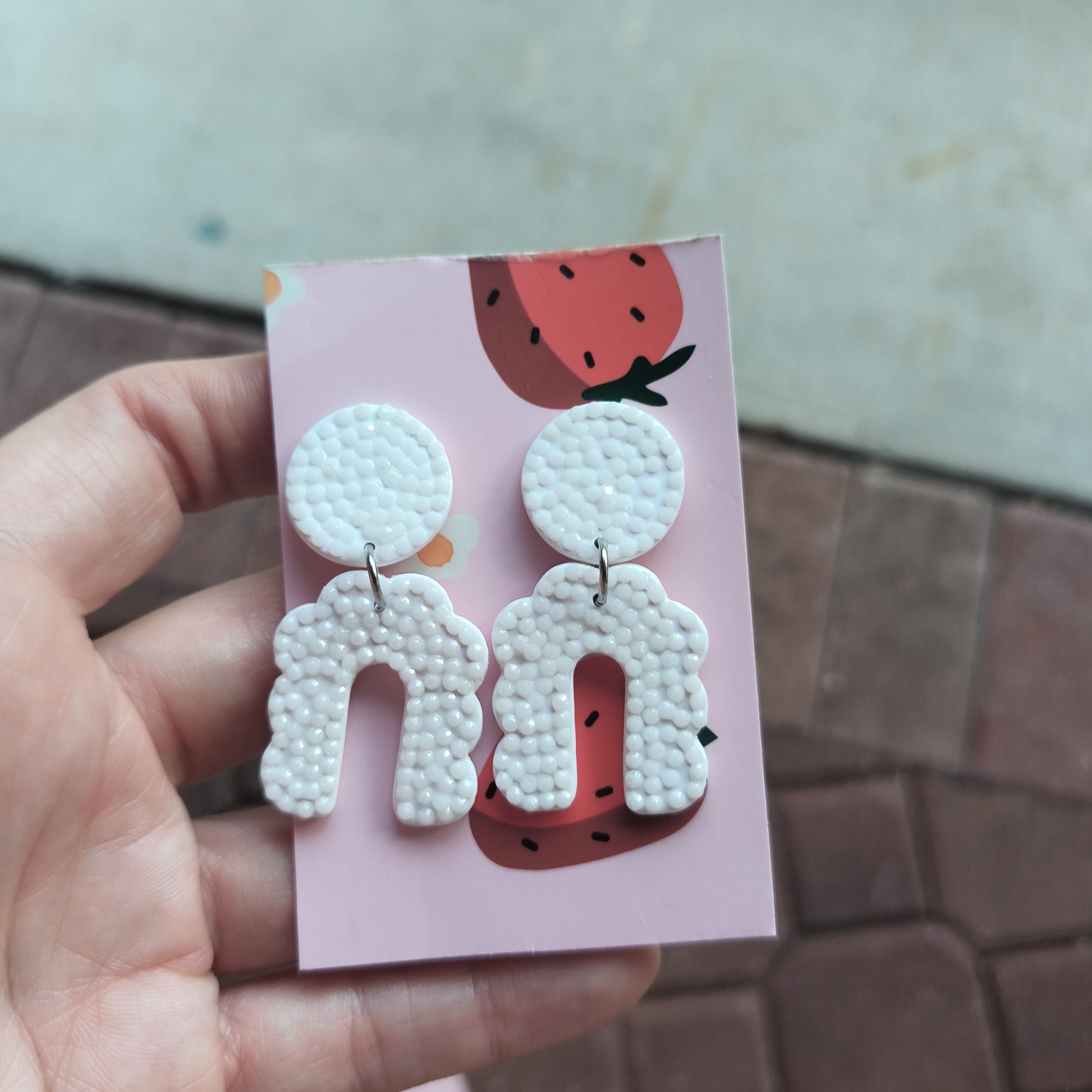 A pair of white, sparkly, modern arch-shaped dangle earrings displayed on a pink card with a strawberry illustration.