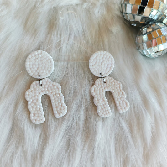 A pair of white, arch-shaped, sparkly earrings with a textured surface displayed on a furry surface, with disco balls in the background.