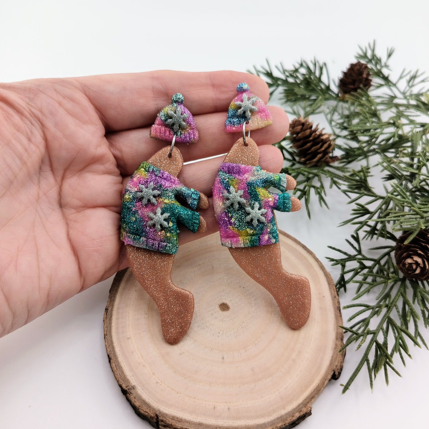 Hand holding Christmas-themed manatee earrings with colorful sweaters and snowflakes on a wooden slice and greenery background.